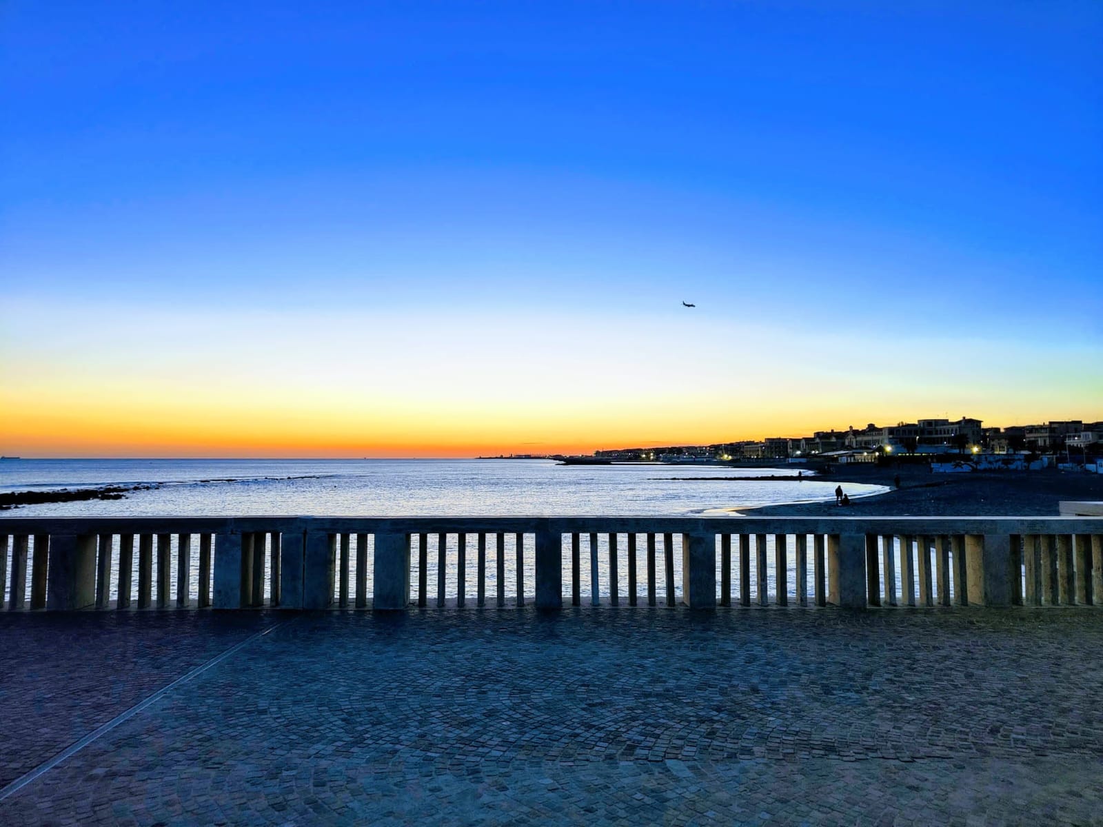 Sunset over the Mediterranean Sea, viewed from the Pontile di Ostia, with the coastline and waterfront buildings in the distance.